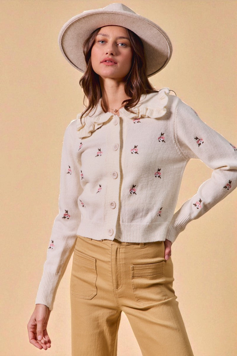 Woman wearing a white embroidered cardigan and beige pants against a beige background