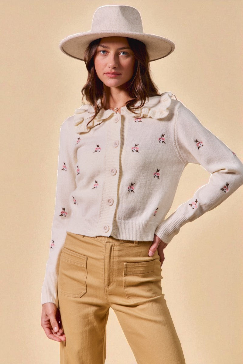 Woman wearing a white embroidered cardigan and beige pants against a beige background