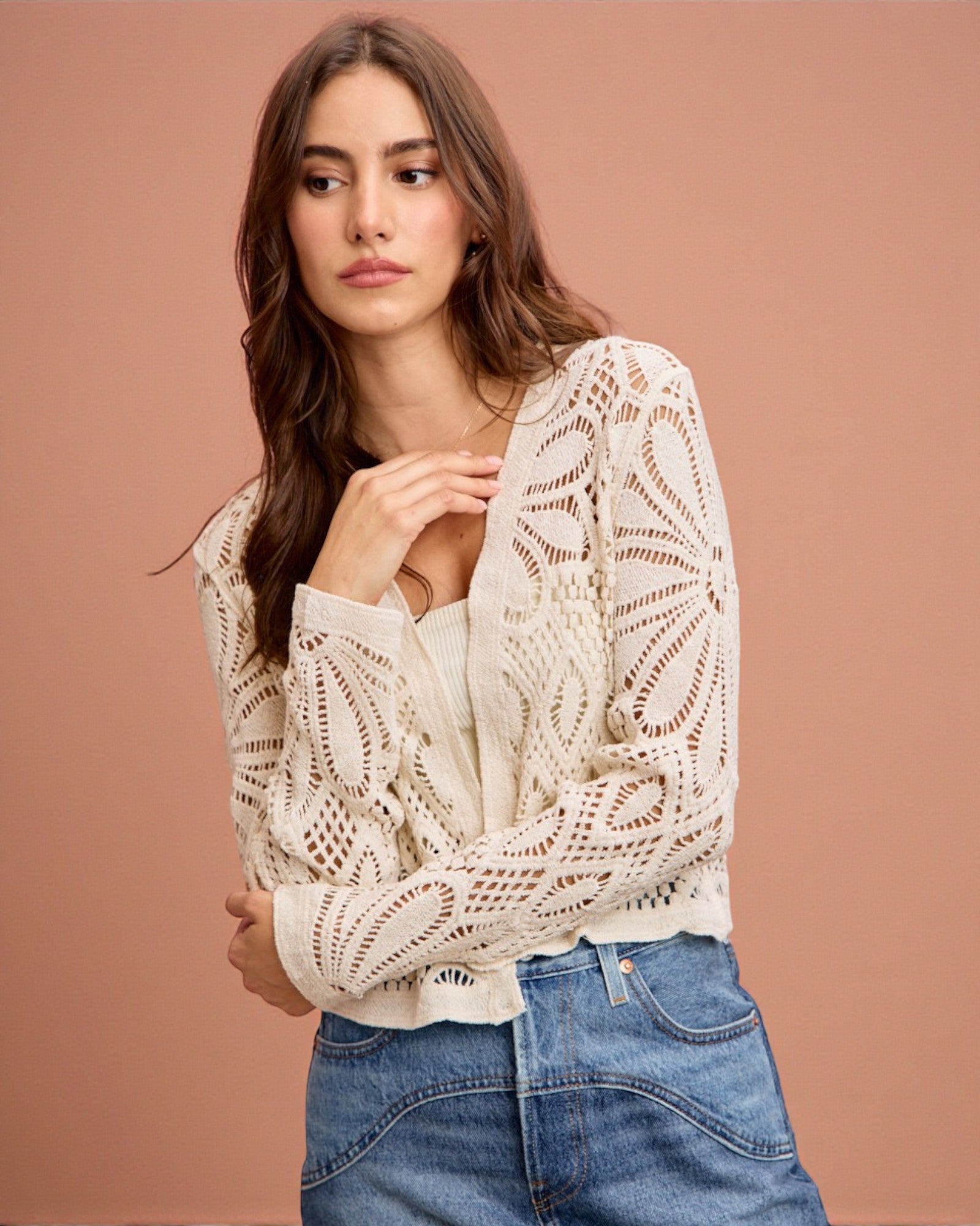 Woman wearing a beige lace top and blue jeans against a brown background
