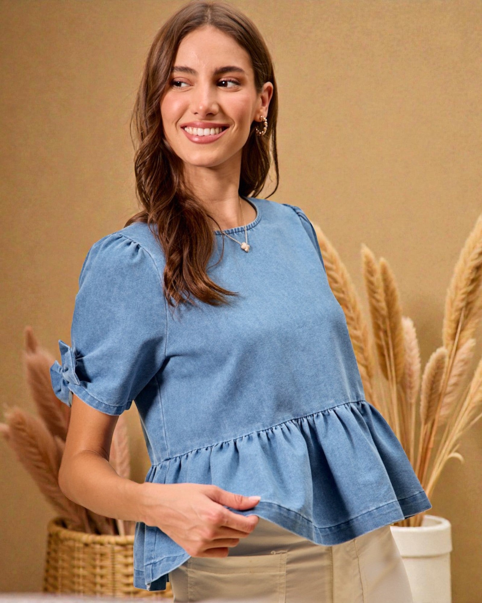 Woman wearing a blue denim top with ruffled details, standing against a beige background with decorative plants.