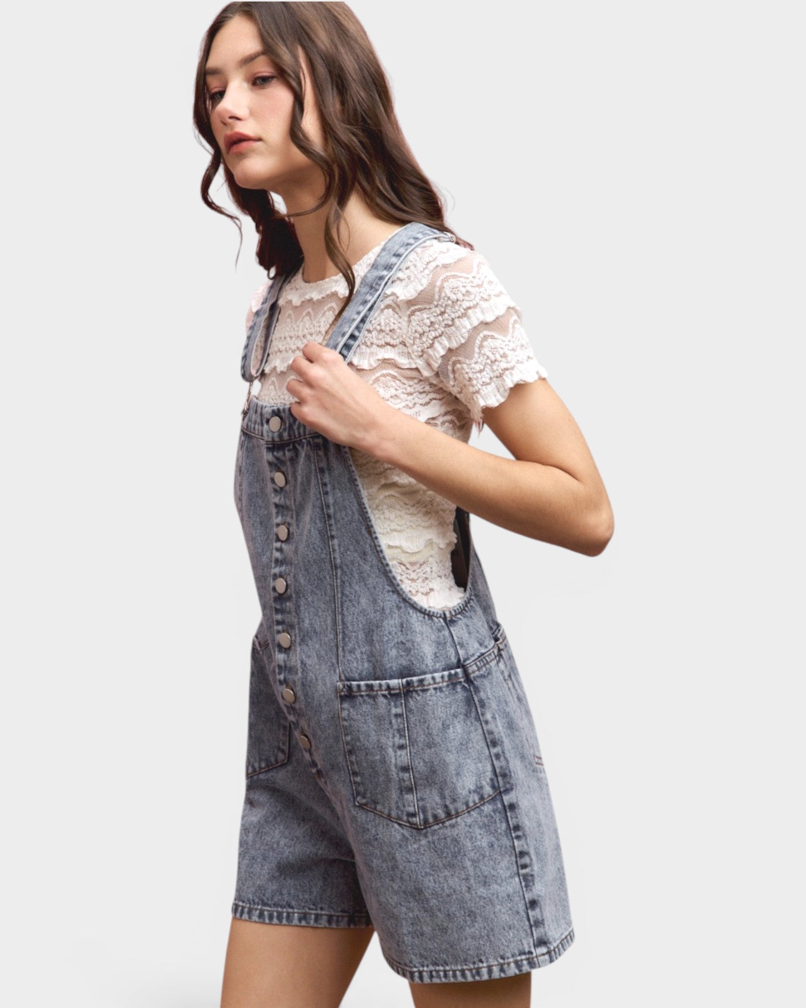 Woman wearing denim overalls over a lace top on a plain background