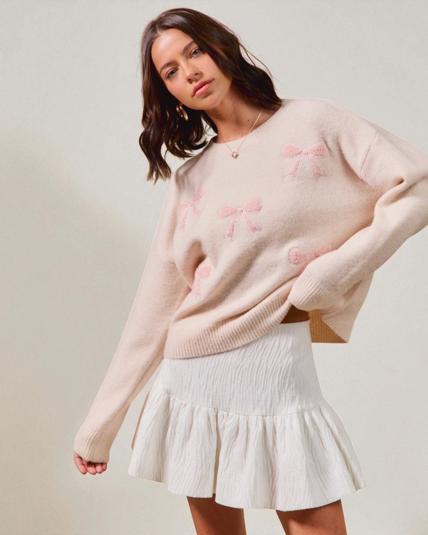 Woman wearing a light pink sweater with floral patterns and a white skirt on a neutral background
