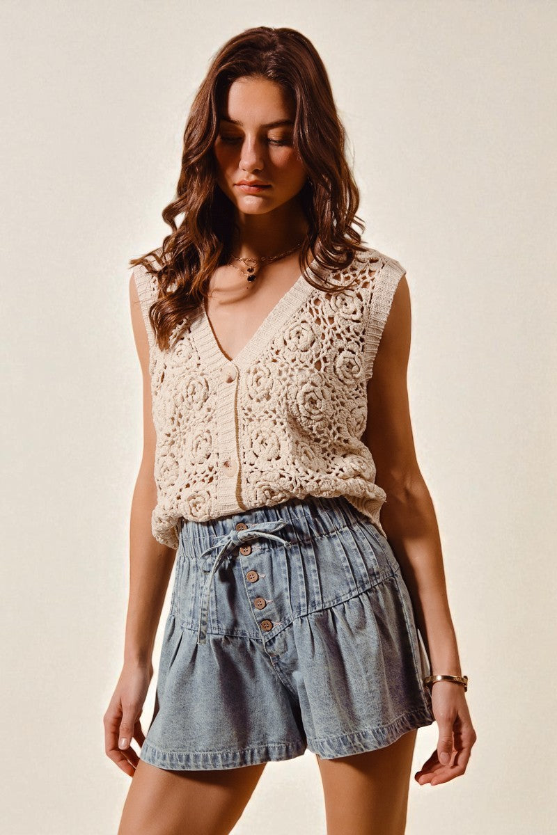 Woman wearing a beige lace top and denim shorts against a plain background