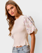 Woman wearing a beige top with floral sleeves and light blue jeans on a plain background