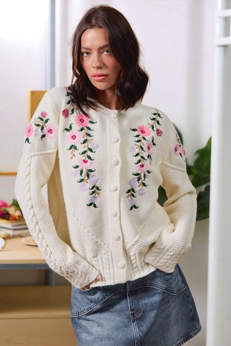 Woman wearing a cream-colored cardigan with floral embroidery in a bright room.