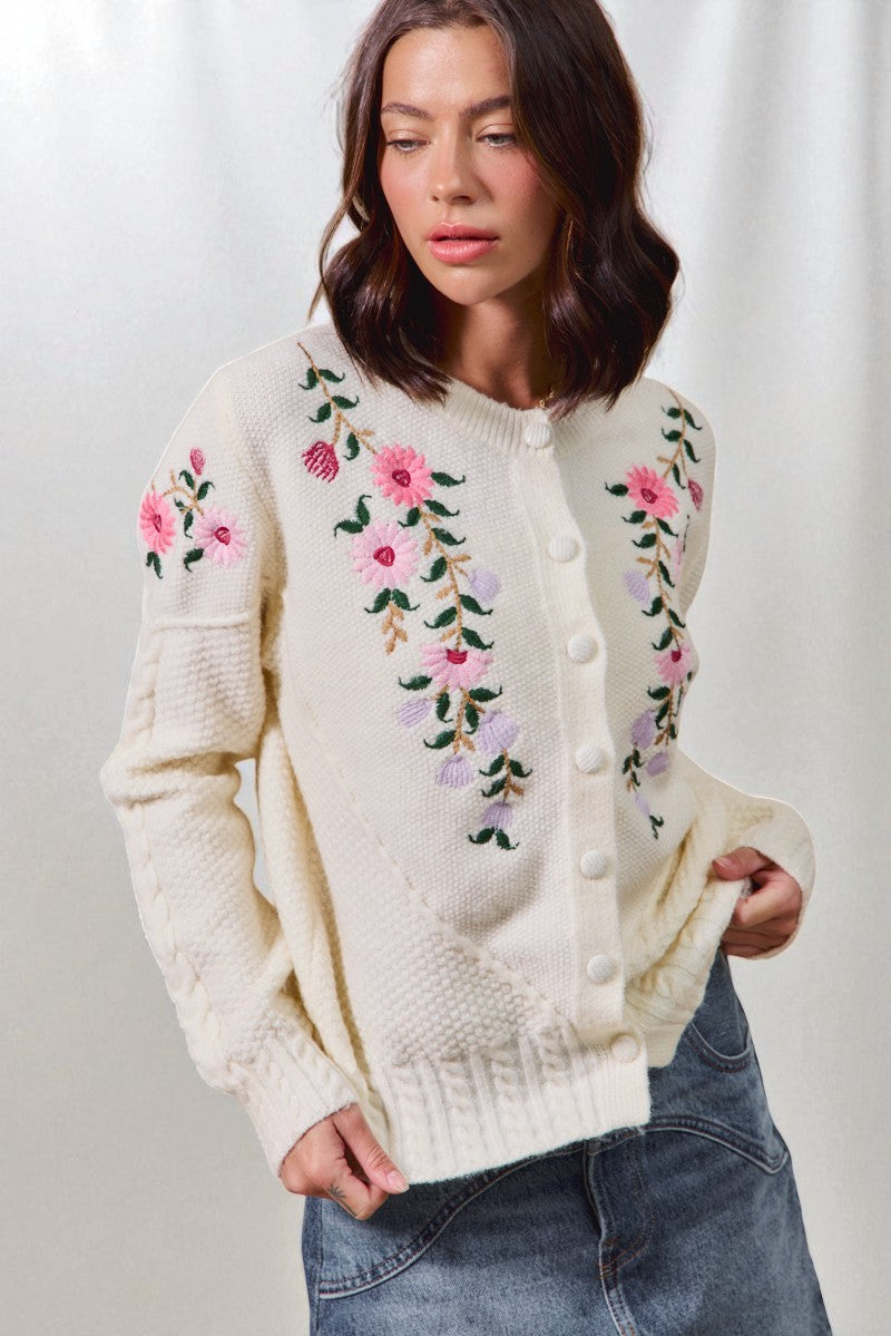 Woman wearing a cream-colored cardigan with floral embroidery against a white background