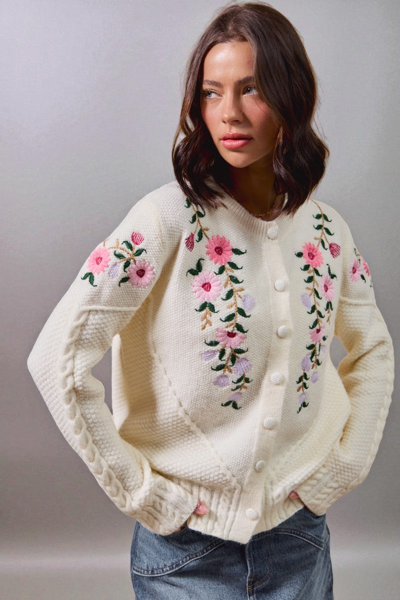 Woman wearing a cream-colored sweater with floral embroidery on a plain background