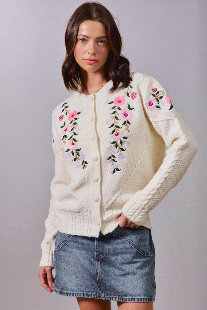 Woman wearing a cream cardigan with floral embroidery against a gray background