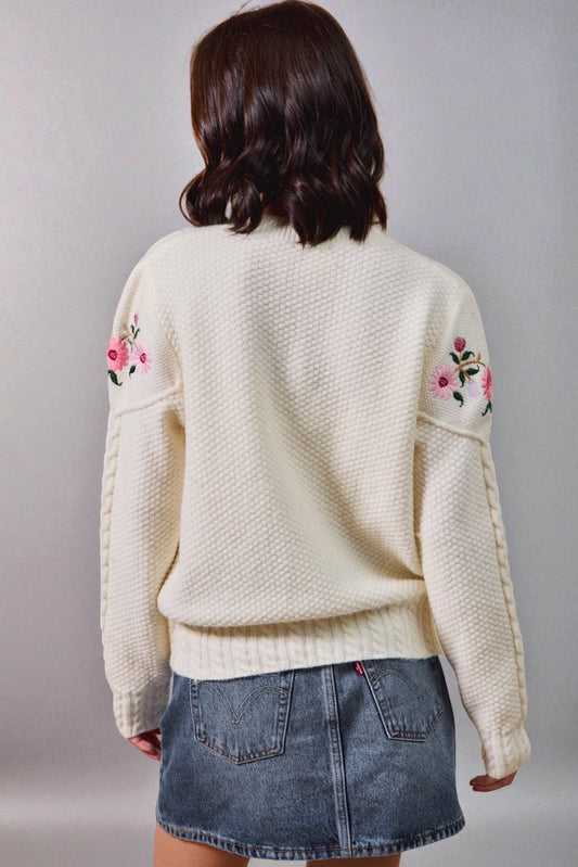 Person wearing a cream sweater with floral embroidery on a plain background