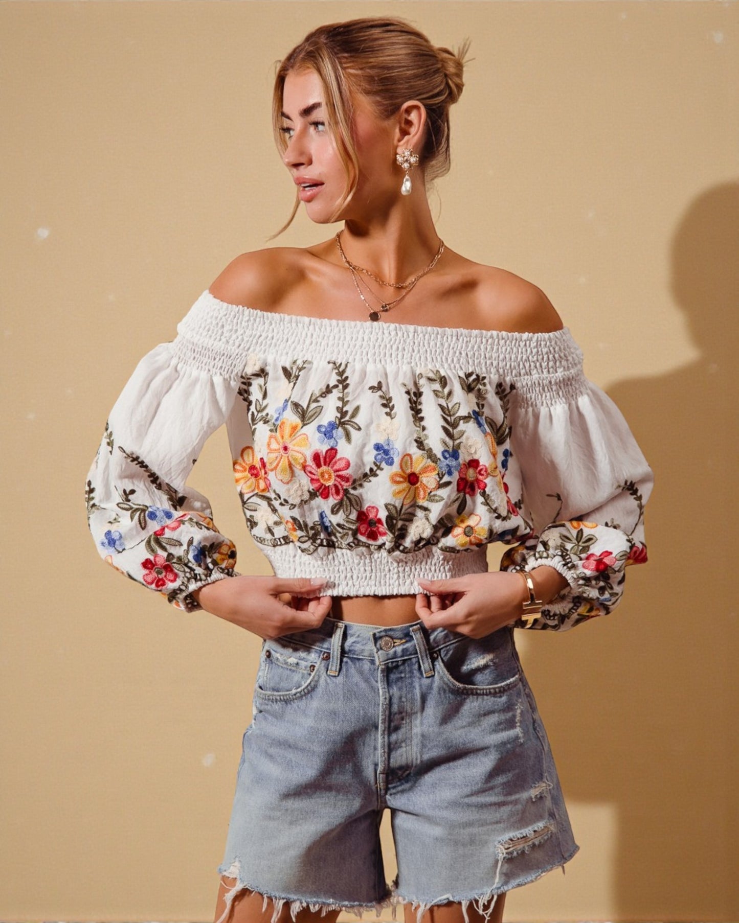Woman wearing a white off-shoulder top with floral embroidery and denim shorts against a beige background