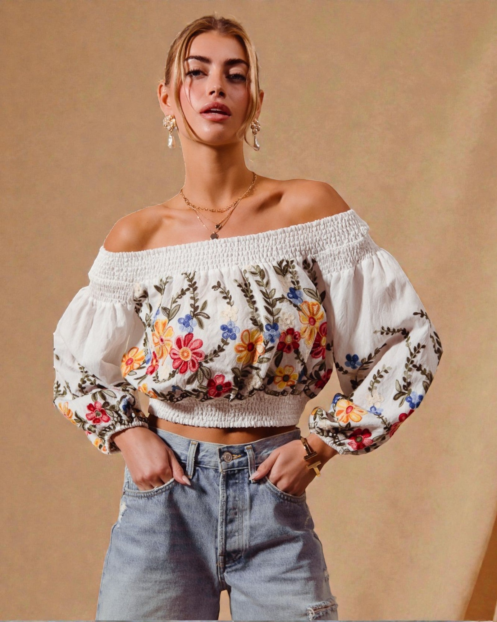 Woman wearing a white off-shoulder top with floral embroidery and high-waisted jeans against a beige background
