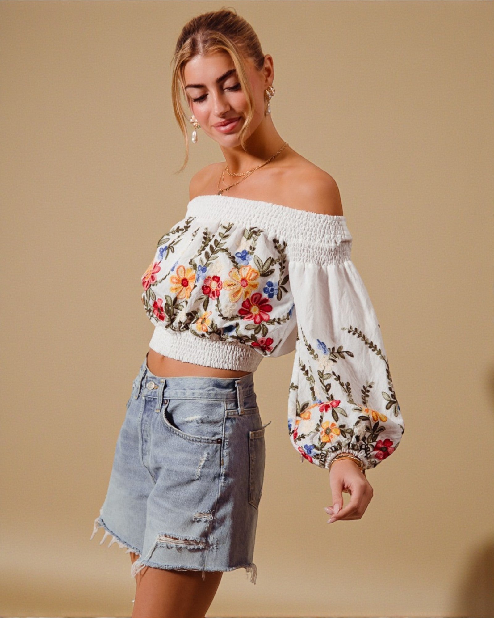Woman wearing a floral embroidered top and denim shorts against a beige background
