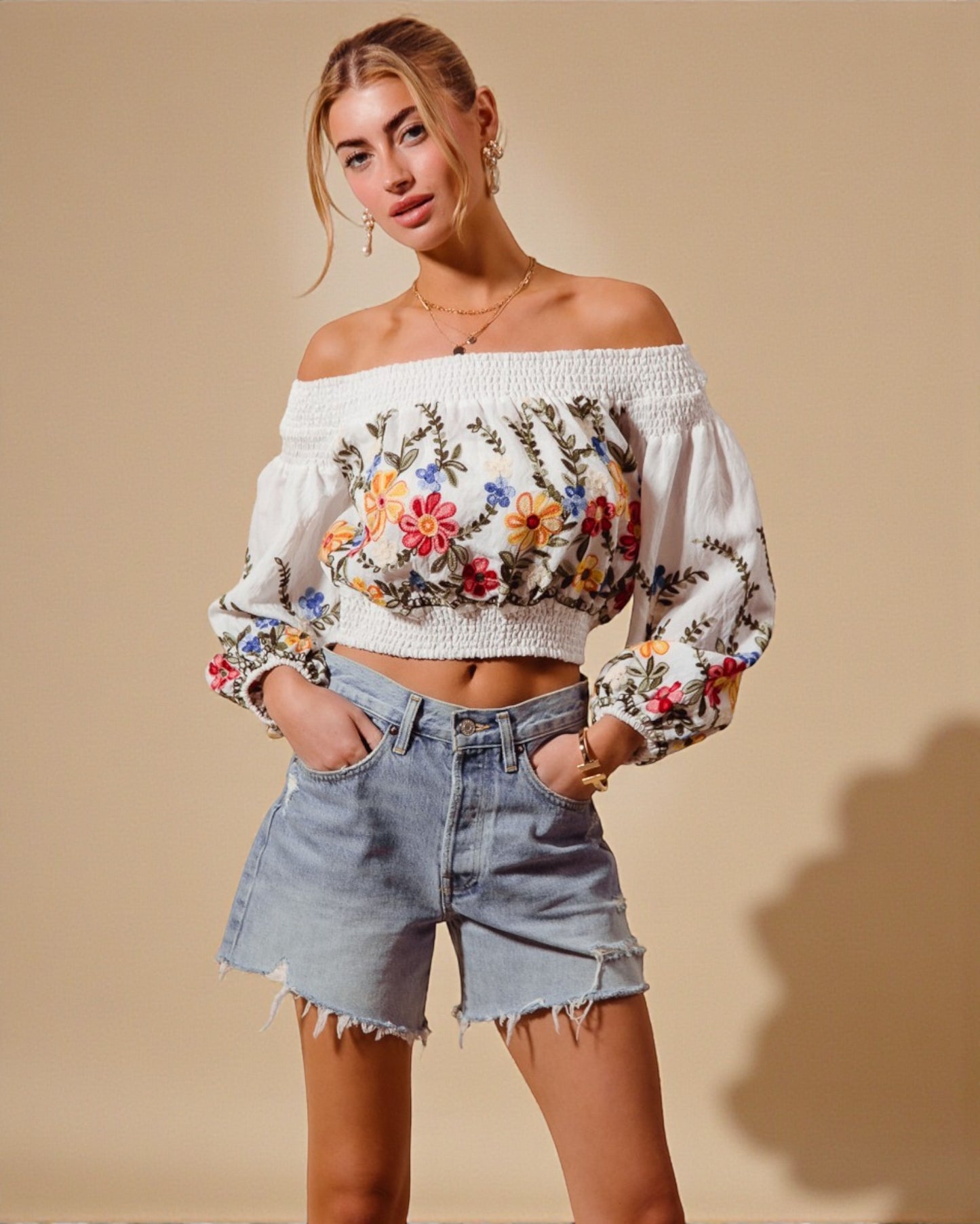 Woman wearing a floral embroidered off-shoulder top and denim shorts on a beige background