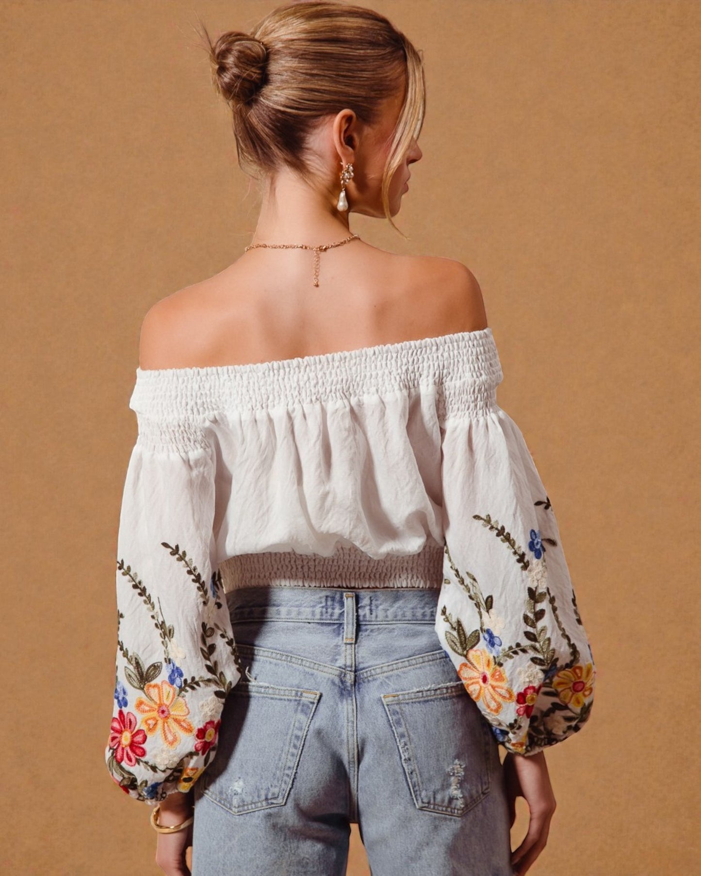 Woman wearing an off-shoulder blouse with floral embroidery and light blue jeans on a beige background