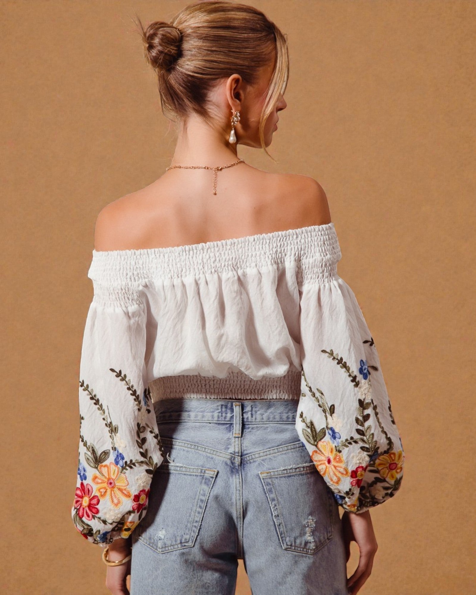 Woman wearing an off-shoulder blouse with floral embroidery and light blue jeans on a beige background