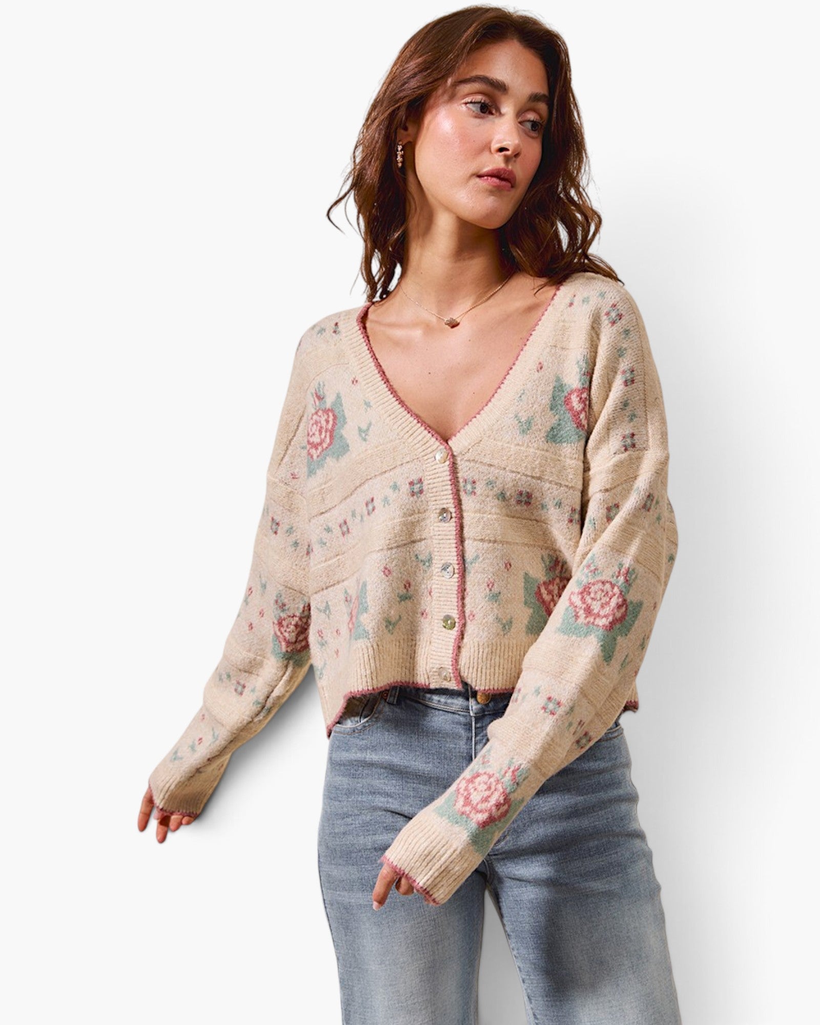 A woman modeling a beige knit cardigan with a pink and green floral pattern, paired with light blue jeans, against a plain white background.