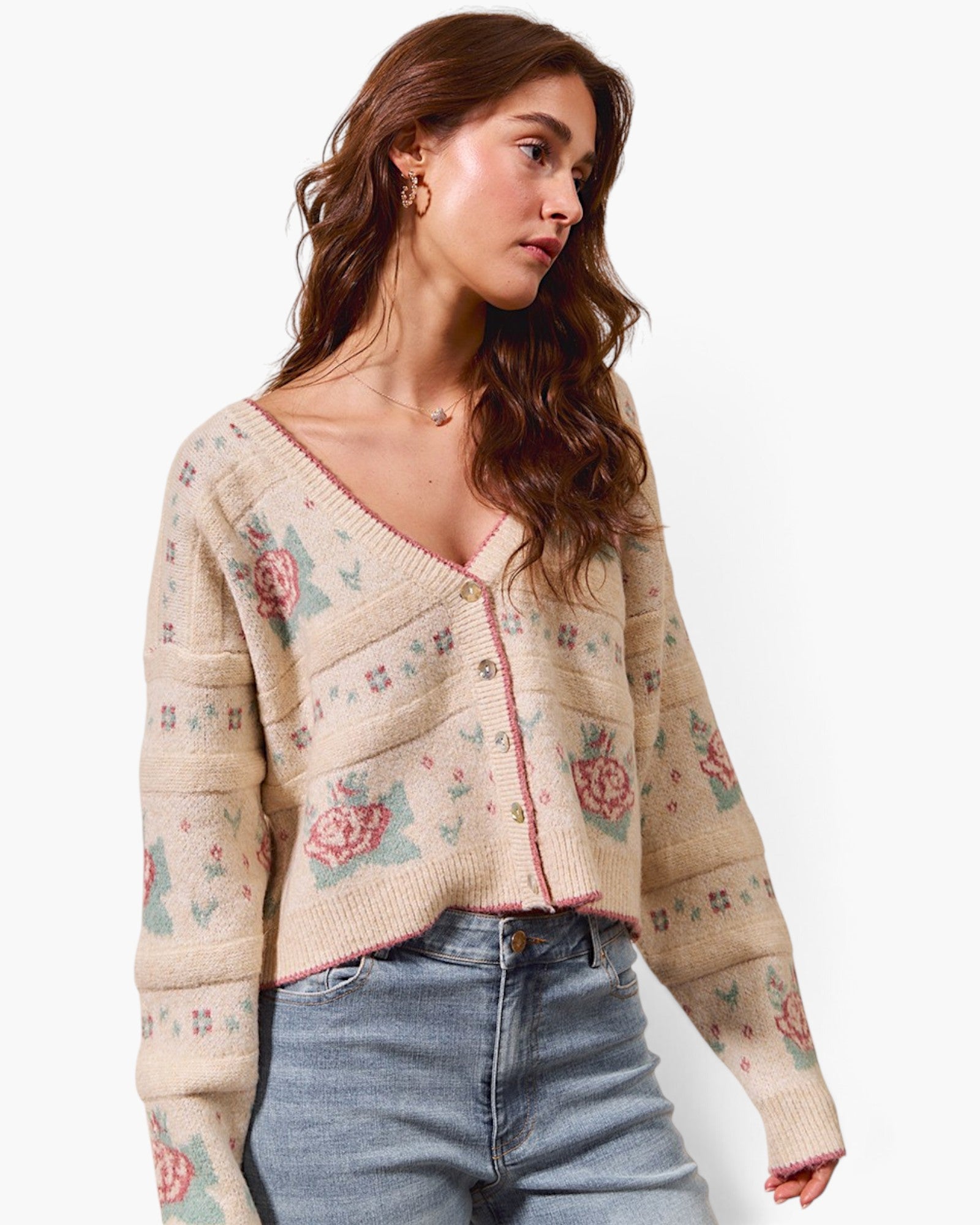 A woman wearing a cream-colored knit cardigan with a pink and green floral pattern, paired with light blue jeans, against a plain white background.