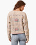 A woman viewed from behind wearing a cream-colored knit sweater with a pink and green floral pattern, paired with light blue jeans, against a plain white background.