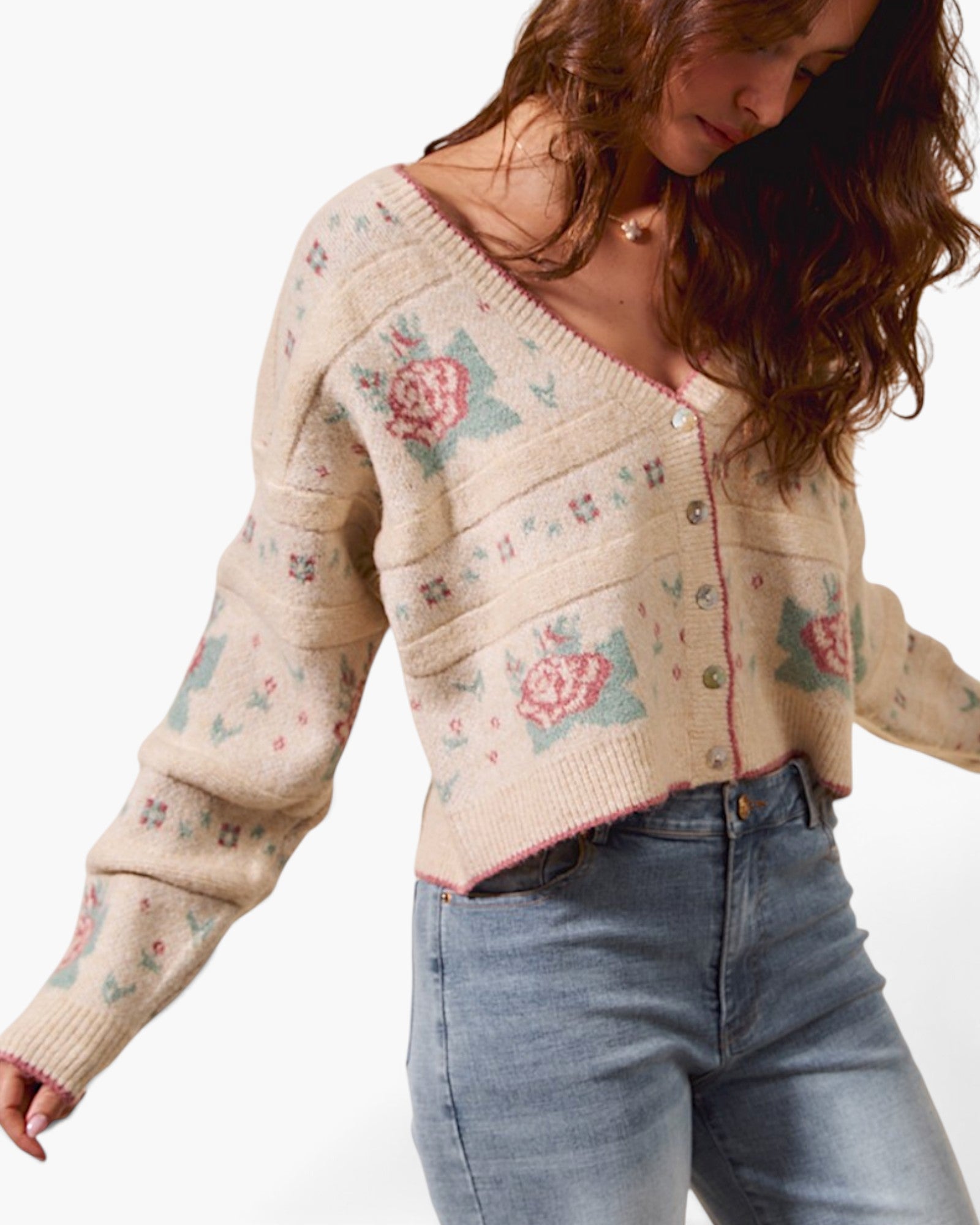 A woman wearing a cream-colored knit cardigan sweater with pink and green floral patterns, paired with blue jeans, against a plain white background.