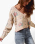 A woman wearing a cream-colored knit cardigan sweater with pink and green floral patterns, paired with blue jeans, against a plain white background.