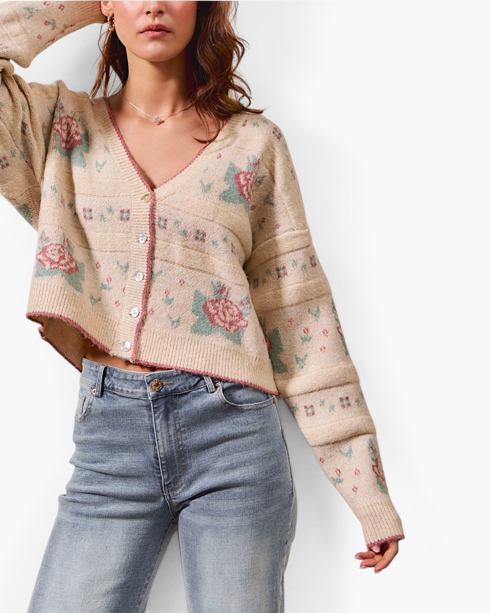 A woman wearing a cropped beige knit cardigan with pink and green floral patterns, paired with light blue jeans, against a plain white background.