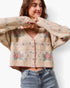 A woman wearing a cropped knit cardigan sweater with a beige base and pink, green, and blue floral patterns, paired with blue jeans against a white studio background.