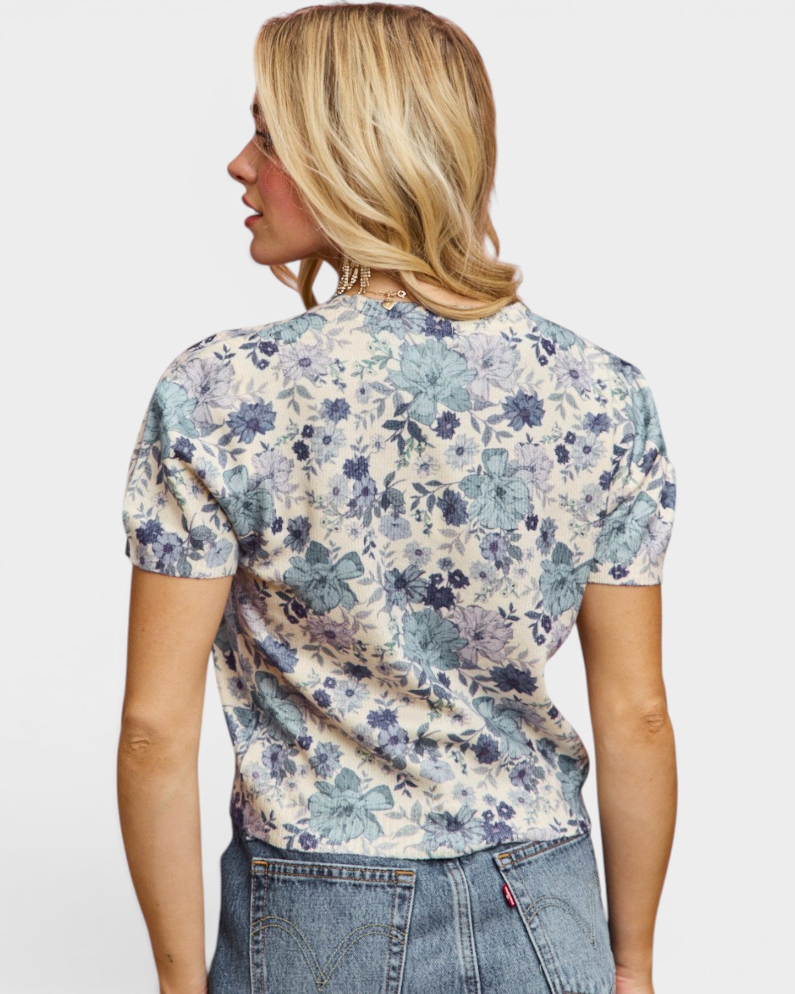 A woman shown from the back wearing a short-sleeve floral t-shirt with a blue and purple pattern on a cream background, paired with blue jeans, against a light gray studio background.