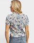 A woman shown from the back wearing a short-sleeve floral t-shirt with a blue and purple pattern on a cream background, paired with blue jeans, against a light gray studio background.
