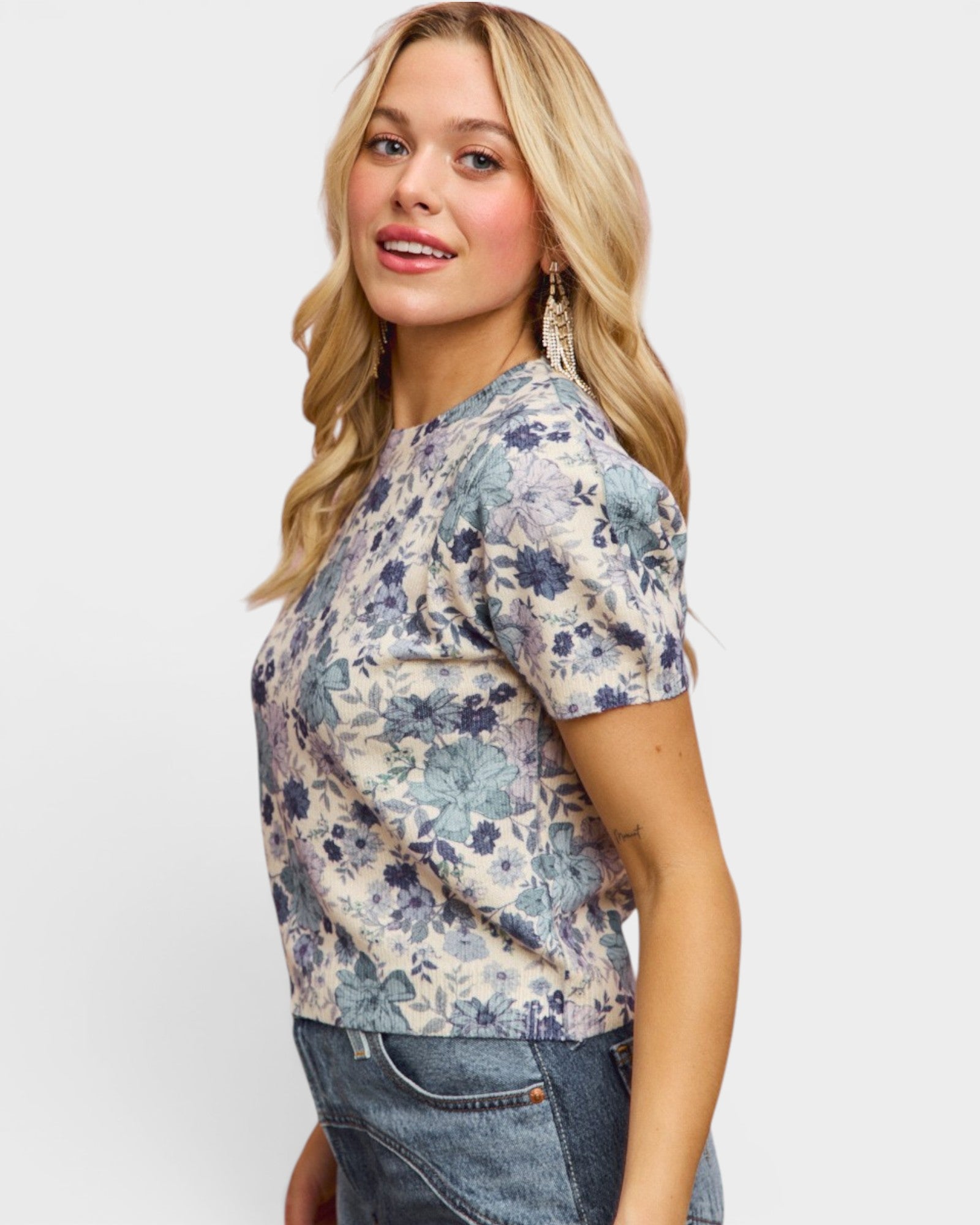 A woman wearing a short-sleeve crewneck top with a blue and beige floral pattern, paired with blue jeans, photographed against a light gray studio background.