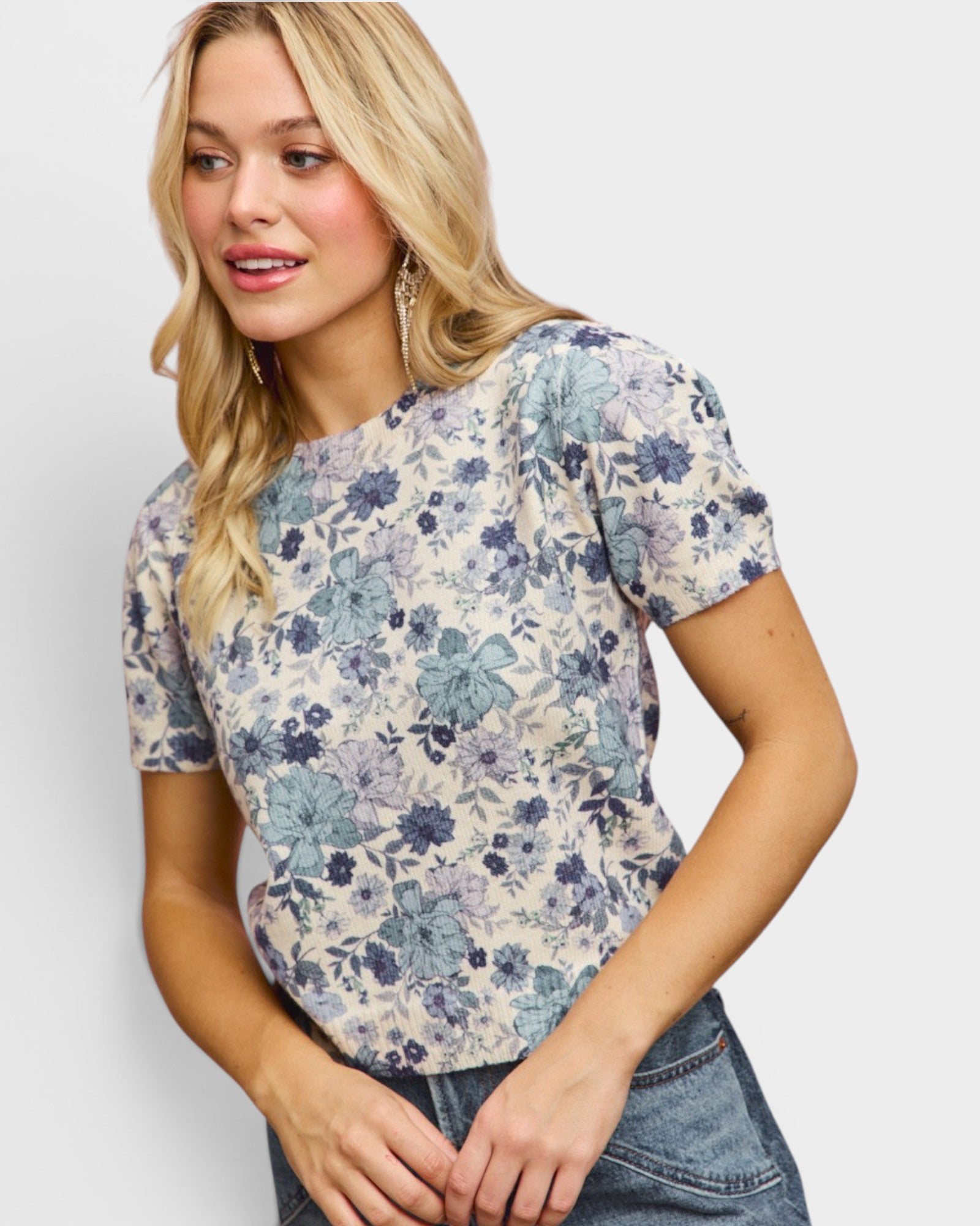 A blonde woman wearing a short-sleeve crewneck top with a blue and gray floral pattern on a cream background, paired with blue jeans, against a light gray studio background.