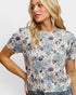 A woman wearing a short-sleeve crewneck top with a blue and purple floral pattern on a cream background, paired with blue jeans and long silver dangle earrings, against a plain light gray background.