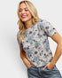 A smiling blonde woman wearing a short-sleeve crewneck top with a blue and purple floral pattern on a cream background, paired with blue jeans, against a light gray studio background.