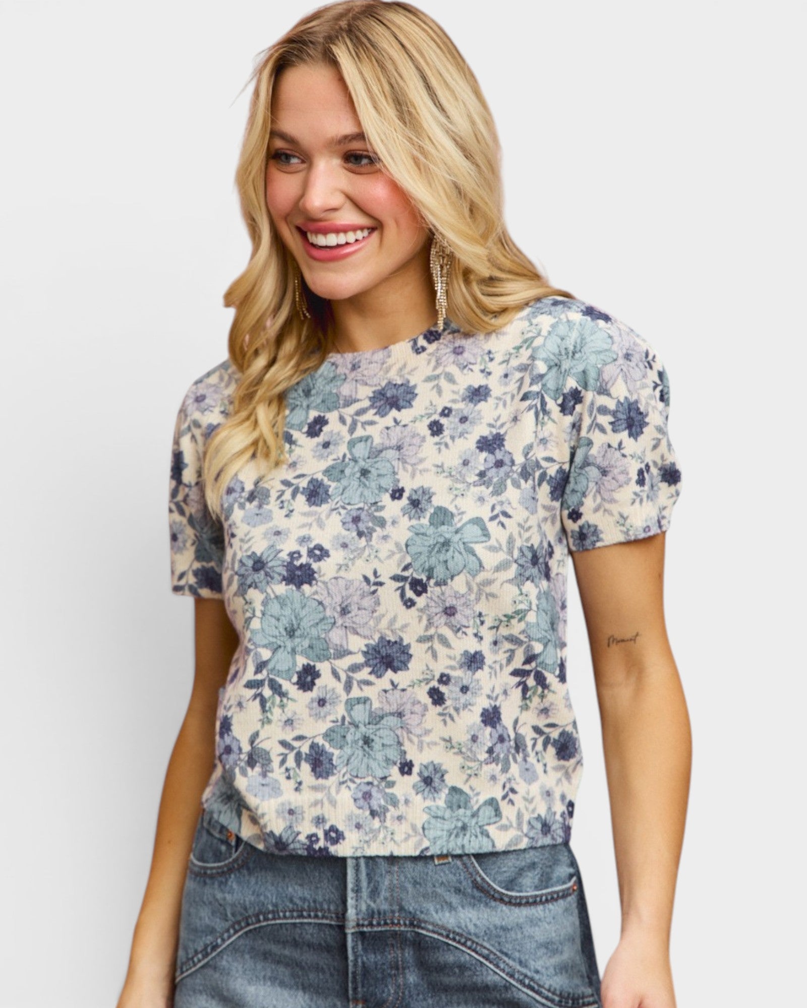 A smiling blonde woman wearing a short-sleeve crewneck top with a blue and purple floral pattern on a cream background, paired with blue jeans, against a light gray studio background.