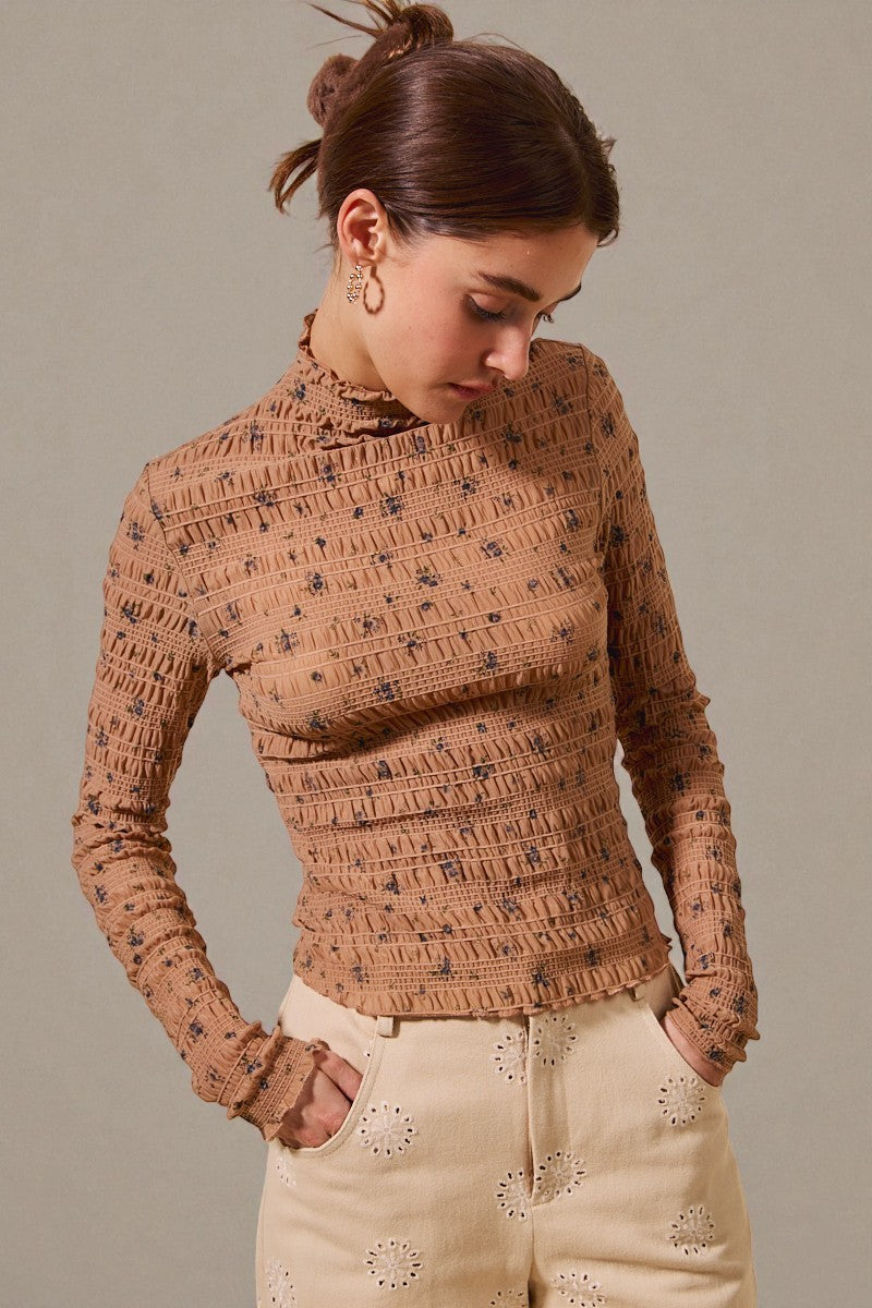 Woman wearing a patterned brown top and beige pants against a neutral background