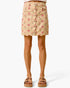A beige denim mini skirt with a pink and green floral print, scalloped hem, and front button closure, shown on a model from the waist down wearing brown platform sandals.
