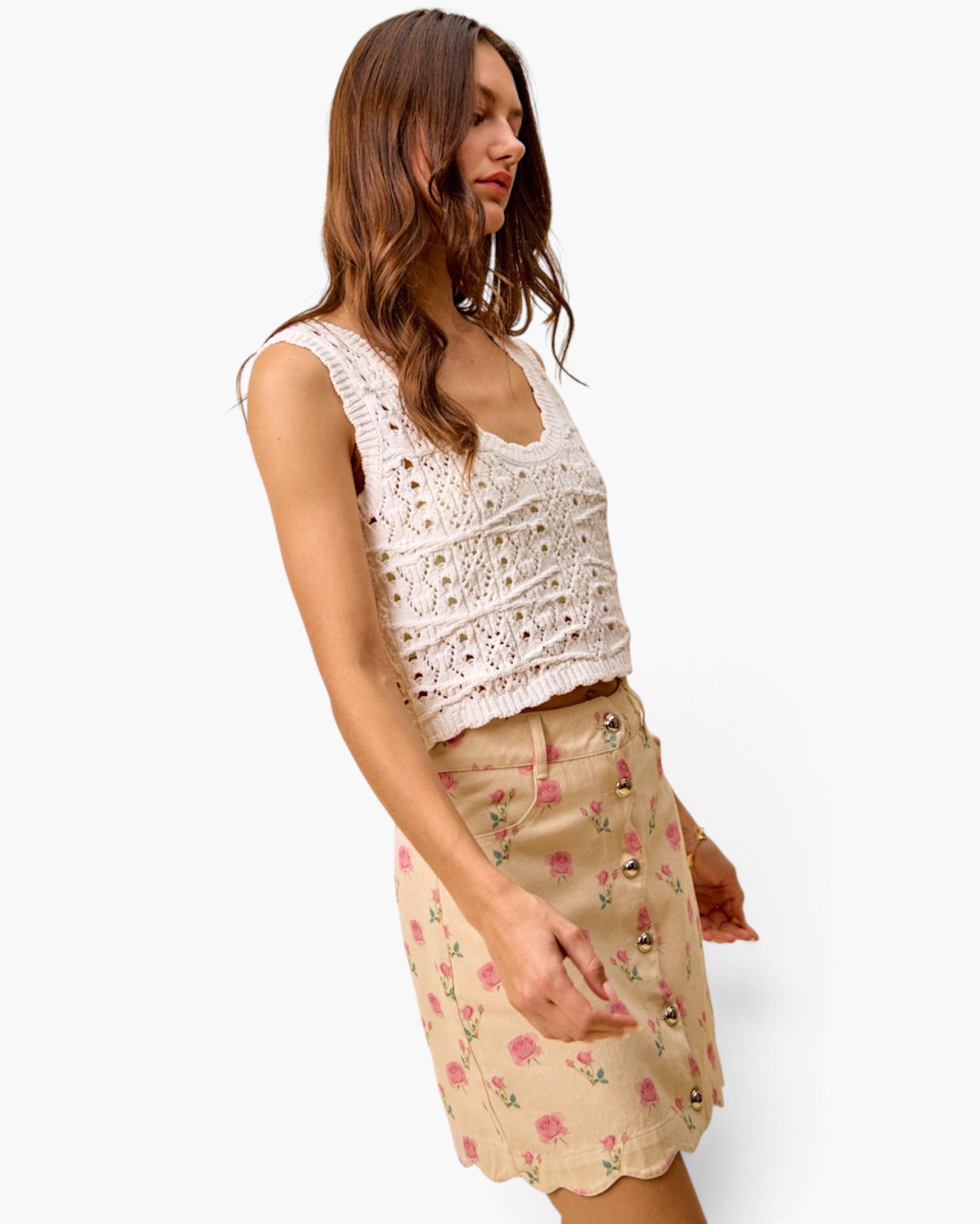 A woman modeling a sleeveless cream crochet knit top paired with a beige mini skirt featuring pink floral prints and scalloped edges, set against a plain white background.
