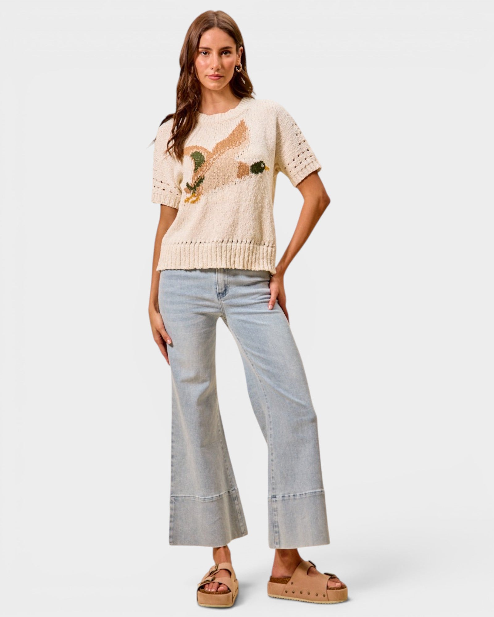 Woman wearing a beige sweater with a bird design and light blue jeans on a white background