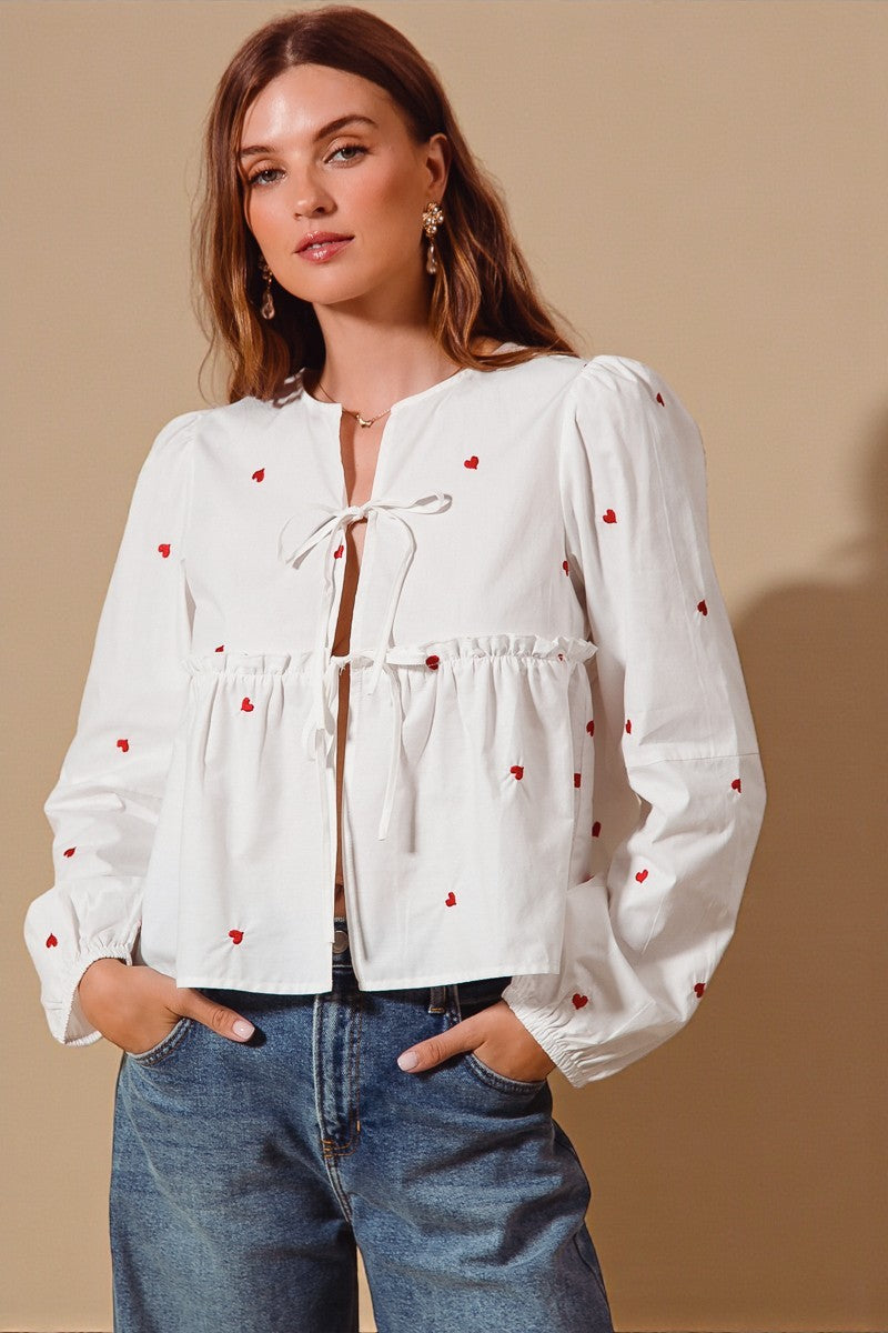 Woman wearing a white blouse with red heart patterns and blue jeans against a beige background