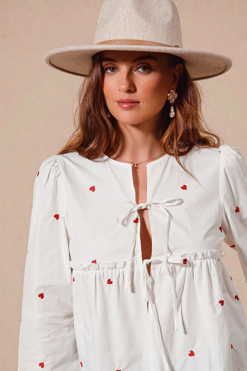 Woman wearing a white dress with red heart patterns and a beige hat against a beige background