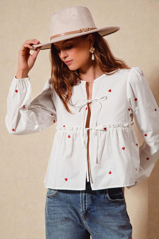 Woman wearing a white blouse with red heart patterns, blue jeans, and a beige hat against a beige background.