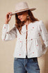 Woman wearing a white blouse with red heart patterns, blue jeans, and a beige hat against a beige background.