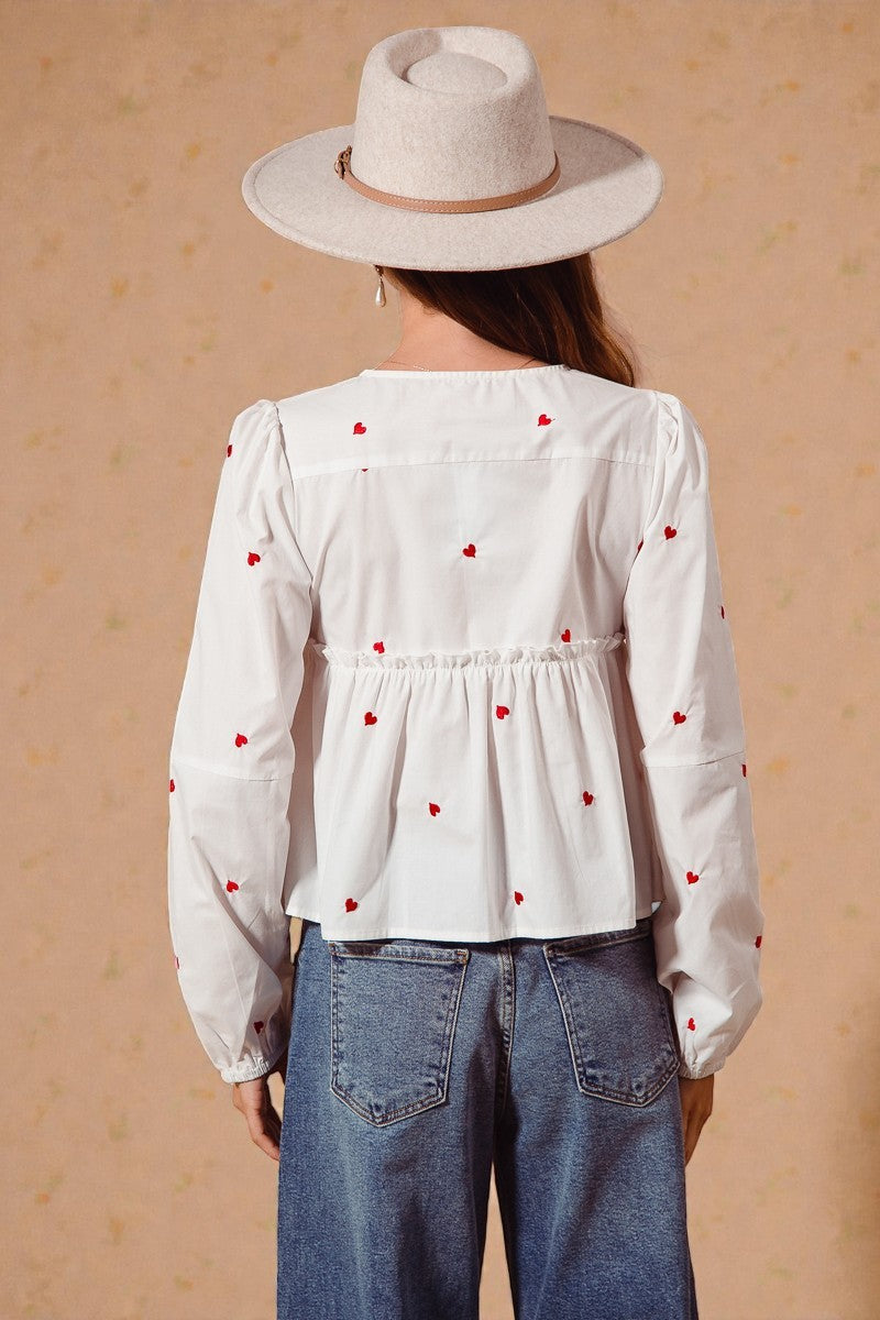 Person wearing a white blouse with red heart patterns and a beige hat on a beige background