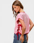Woman wearing a pink top with ruffled sleeves and red accents, standing against a plain background.