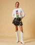 Woman wearing a white sweater with colorful bow designs, a patterned skirt, and white boots on a beige background