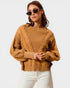 Woman wearing a mustard yellow cable knit sweater and white pants on a plain background