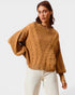 Woman wearing a brown cable knit sweater and white pants on a light gray background