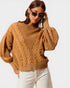 Woman wearing a brown cable knit sweater and white pants on a plain background
