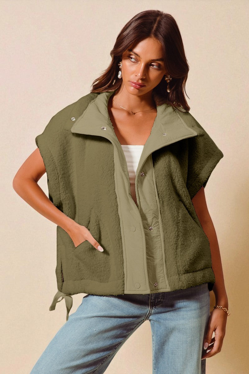 Woman wearing an olive green vest over a white top and blue jeans on a beige background