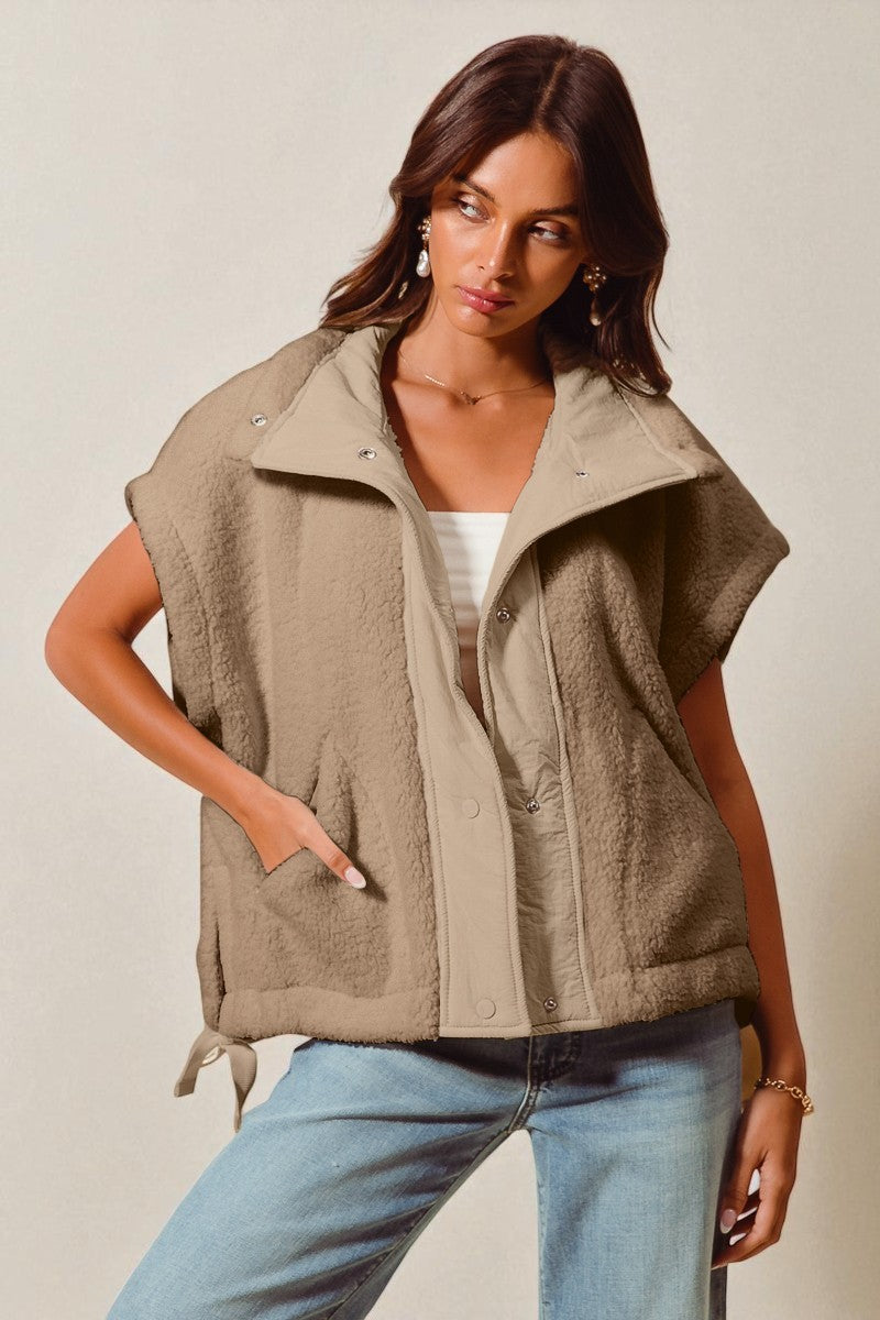 Woman wearing a beige vest over a white top and blue jeans on a plain background