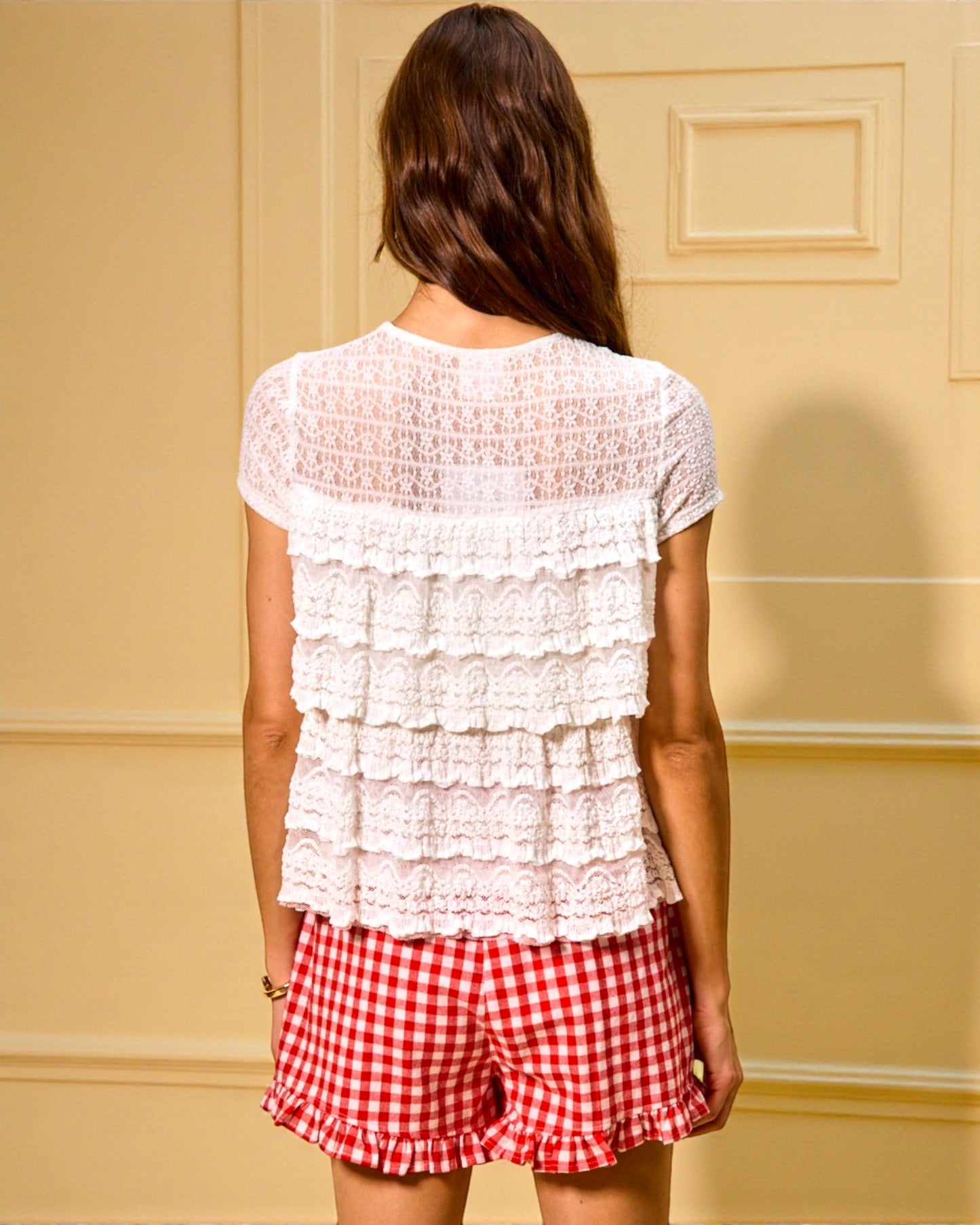 Woman wearing a white ruffled top and red checkered shorts against a beige wall.